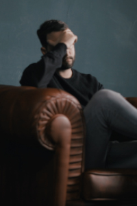 A man sits in an arm chair with his hand covering his face. His expression looks sad and anxious